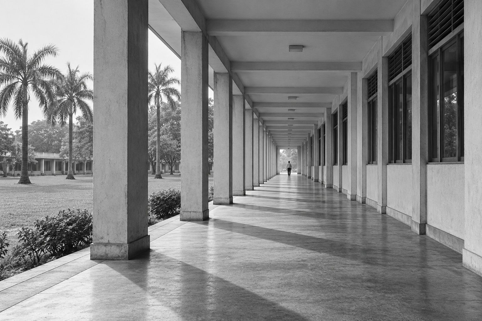 Black and white corridor of square concrete columns throwing stripes of shadow, a tiny figure at the end