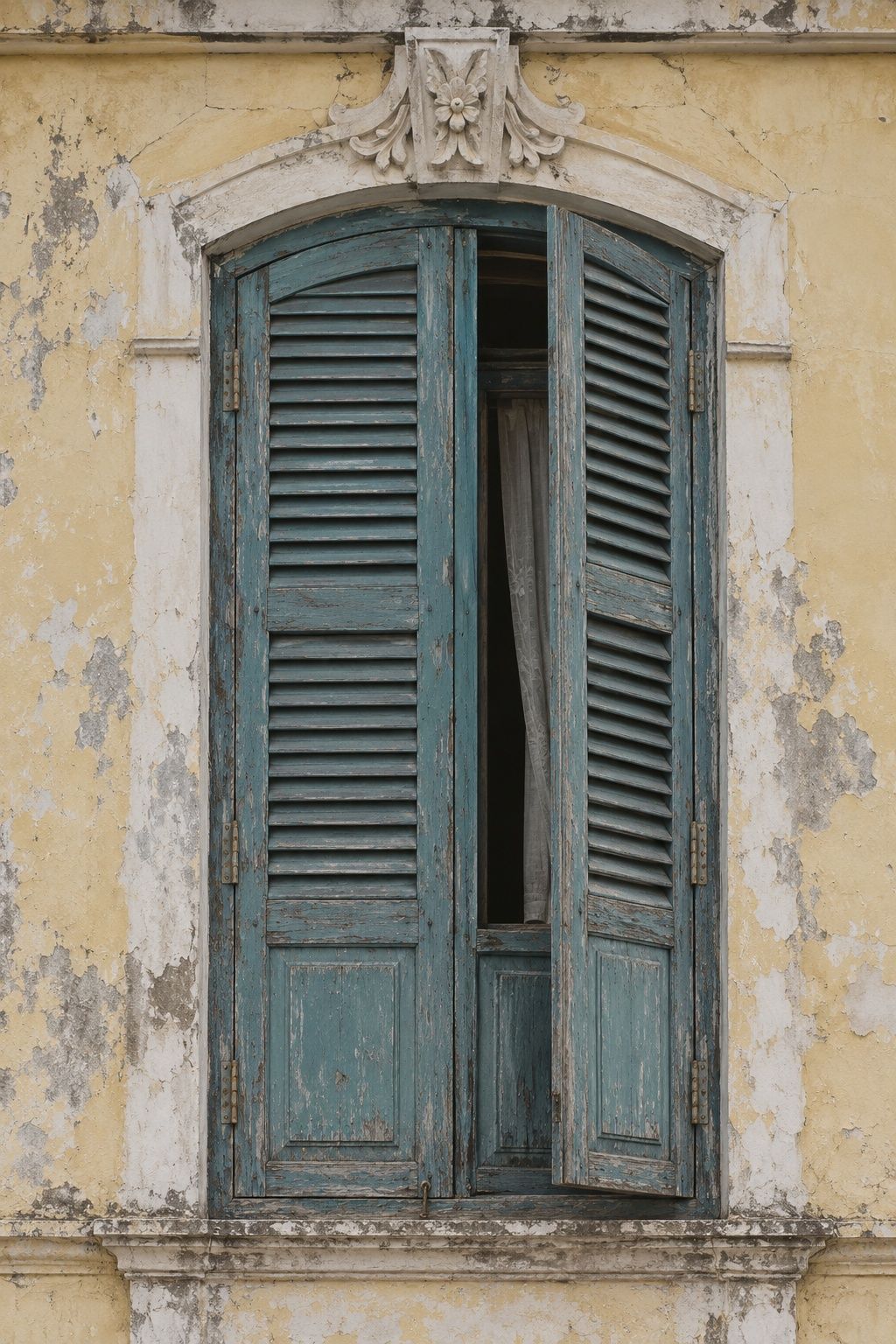 Weathered blue-green timber shutters, one half open, set in a cracked pale yellow wall